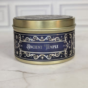 Ancient Temple 8oz soy candle in a gold tin with a navy and gold foil label, on white marble