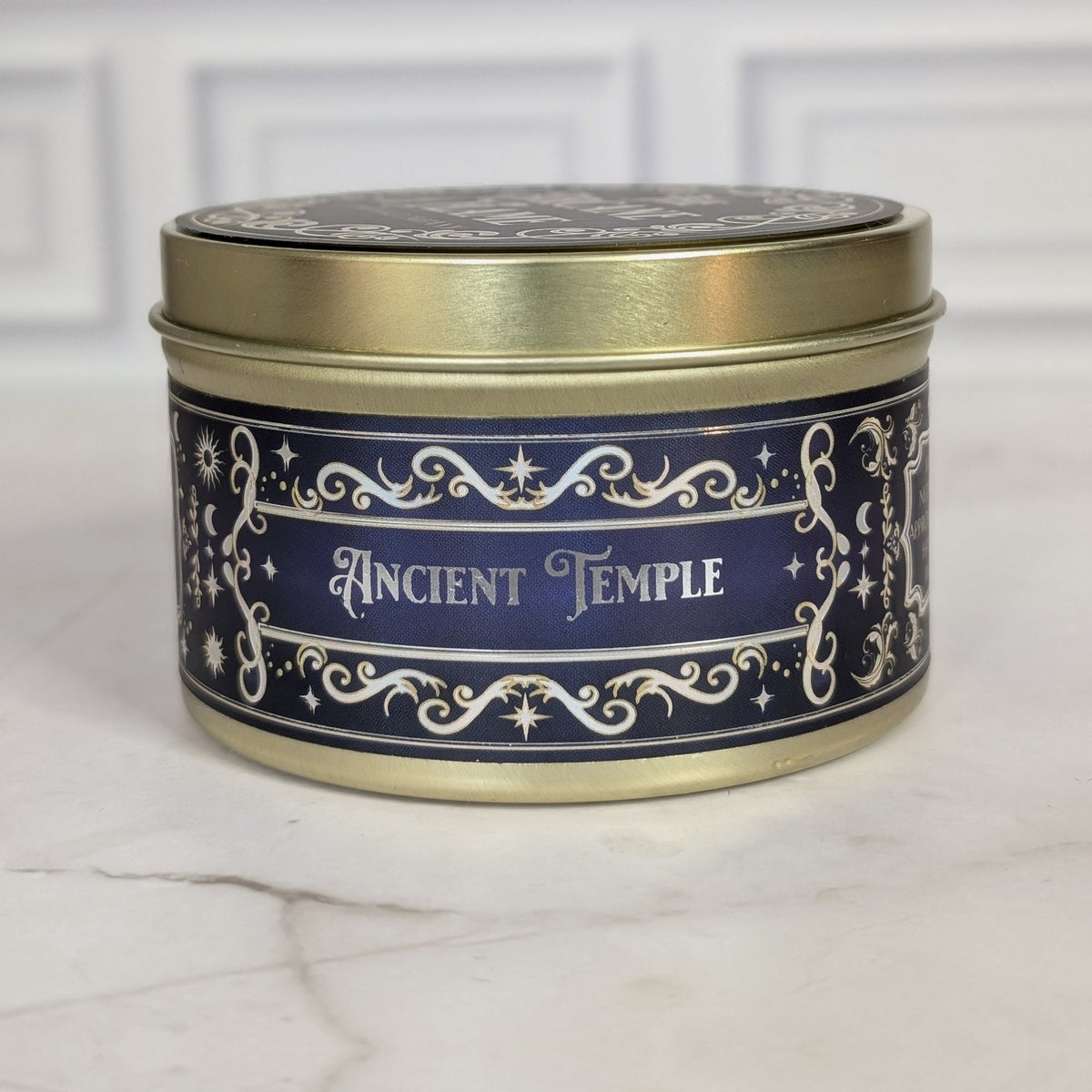 Ancient Temple 8oz soy candle in a gold tin with a navy and gold foil label, on white marble