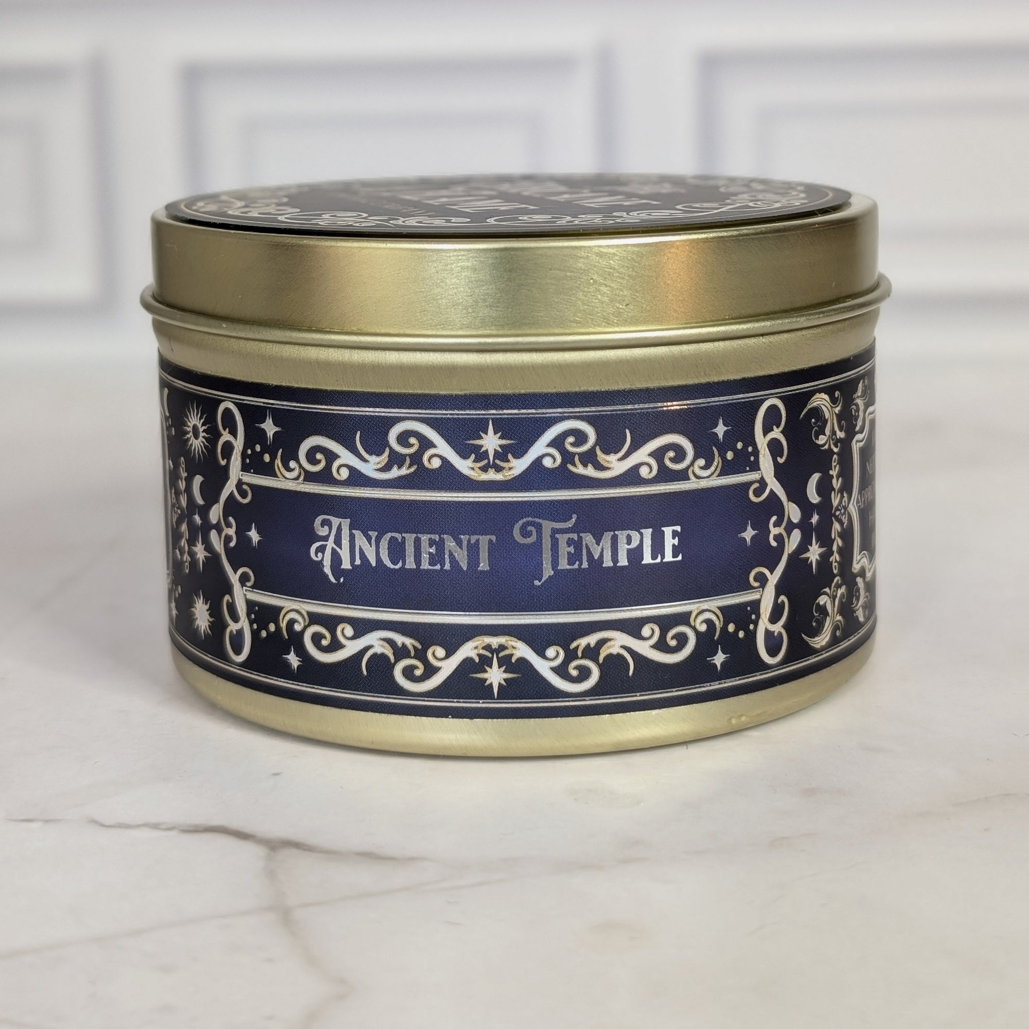Ancient Temple 8oz soy candle in a gold tin with a navy and gold foil label, on white marble
