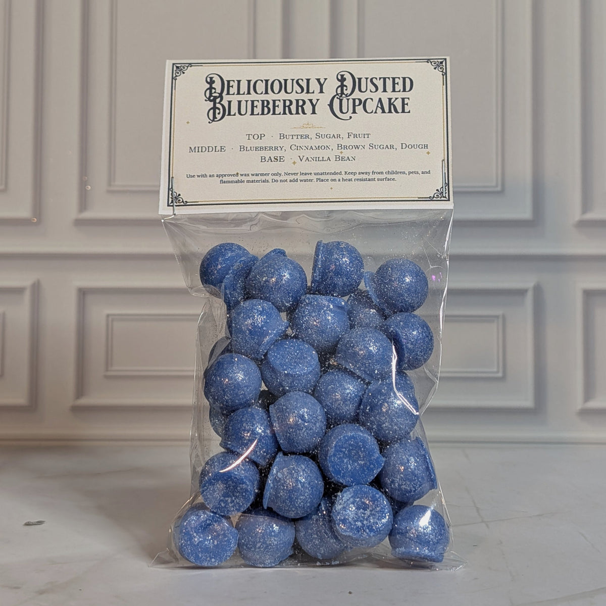 Deliciously Dusted Blueberry Cupcake wax melt drops in a sealed cello bag with a navy and gold topper card, on white marble