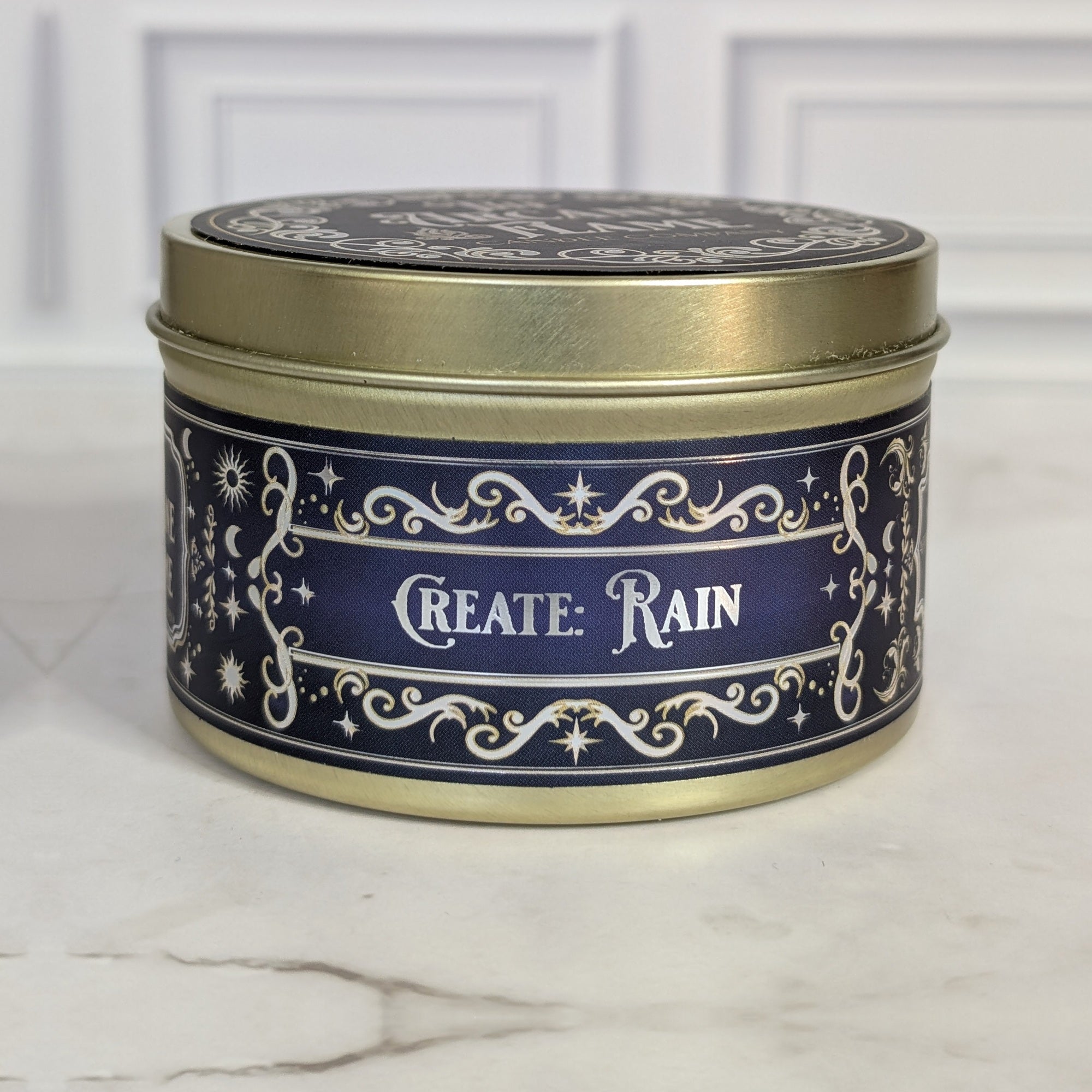Create: Rain 8oz soy candle in a gold tin with a navy and gold foil label, on white marble