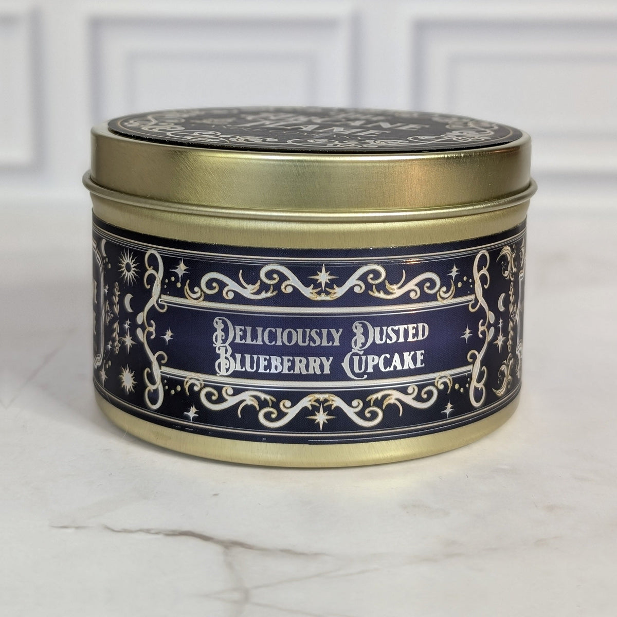 Deliciously Dusted Blueberry Cupcake 8oz soy candle in a gold tin with a navy and gold foil label, on white marble
