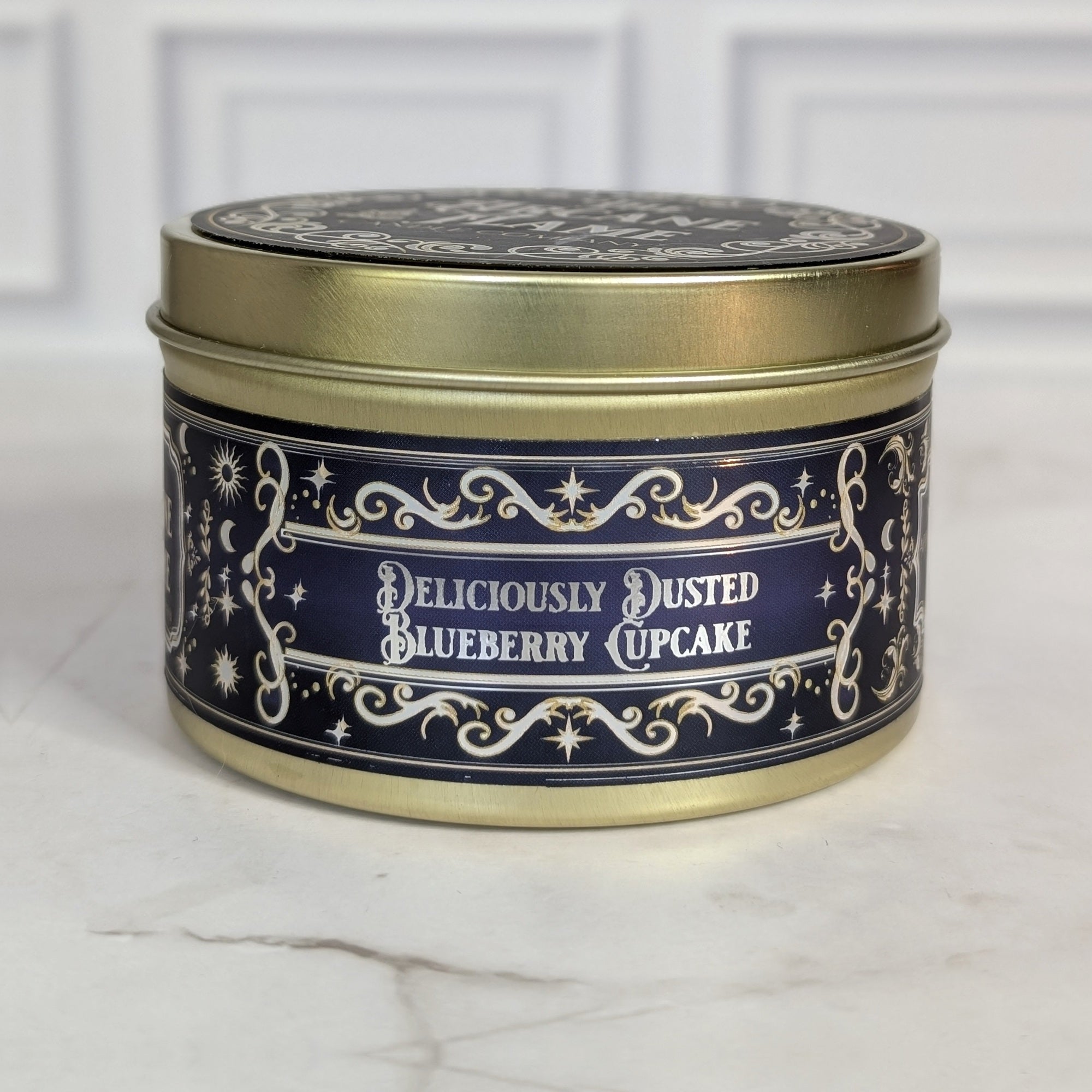 Deliciously Dusted Blueberry Cupcake 8oz soy candle in a gold tin with a navy and gold foil label, on white marble