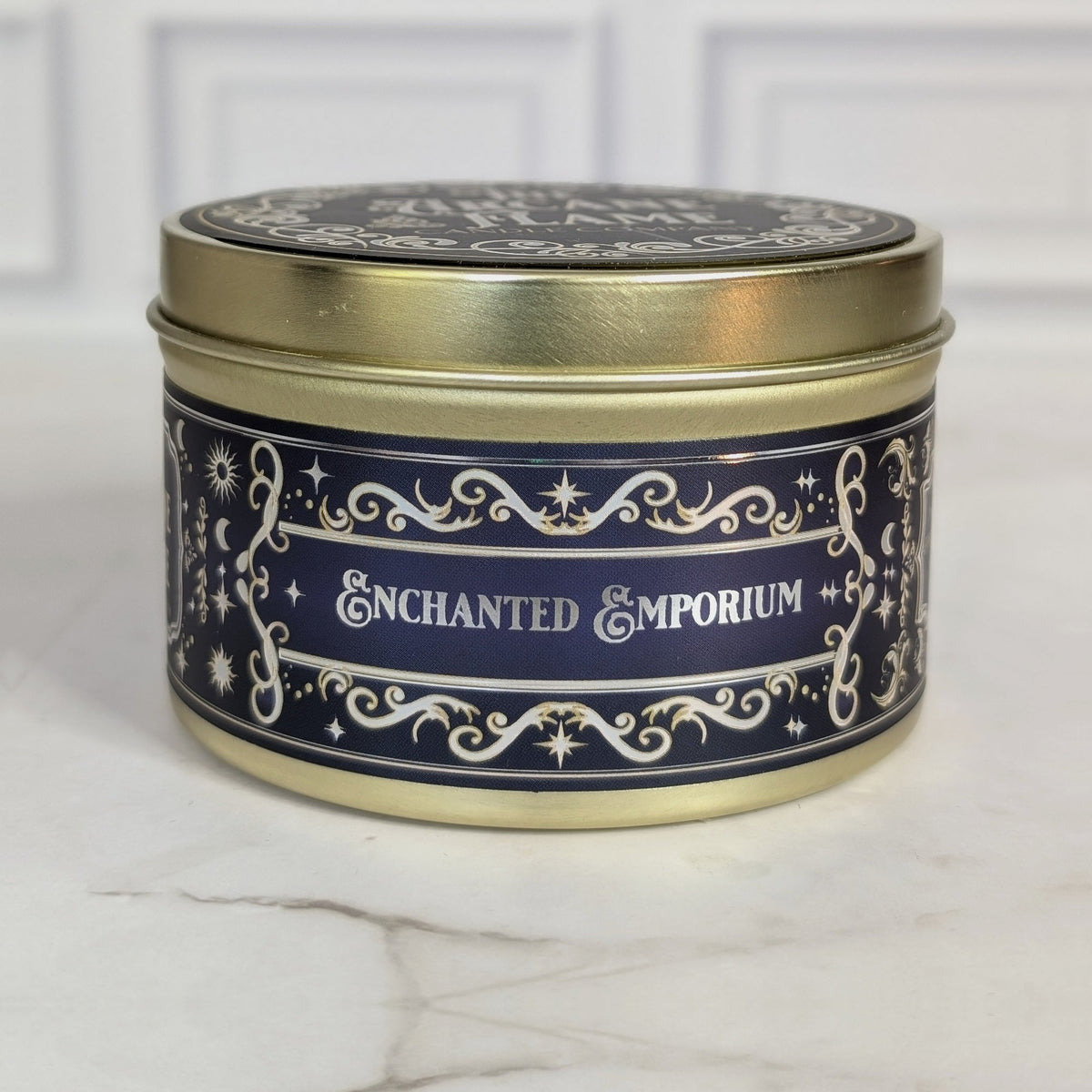 Enchanted Emporium 8oz soy candle in a gold tin with a navy and gold foil label, on white marble