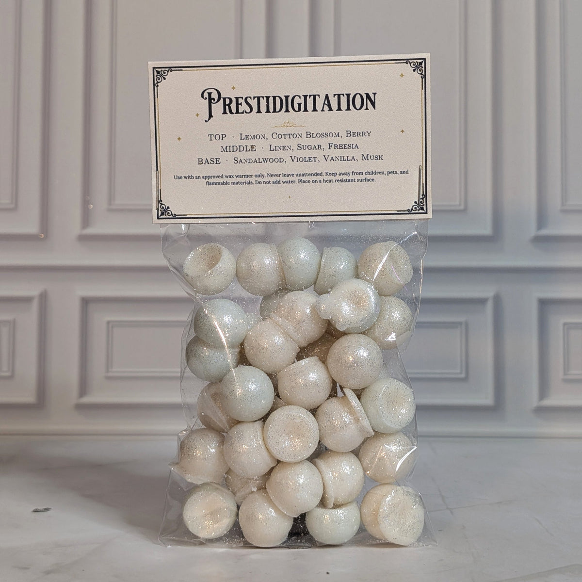 Prestidigitation wax melt drops in a sealed cello bag with a navy and gold topper card, on white marble