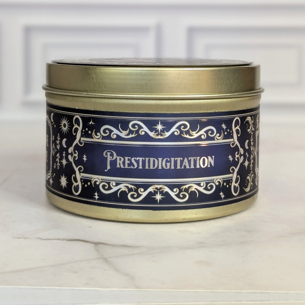Prestidigitation 8oz soy candle in a gold tin with a navy and gold foil label, on white marble